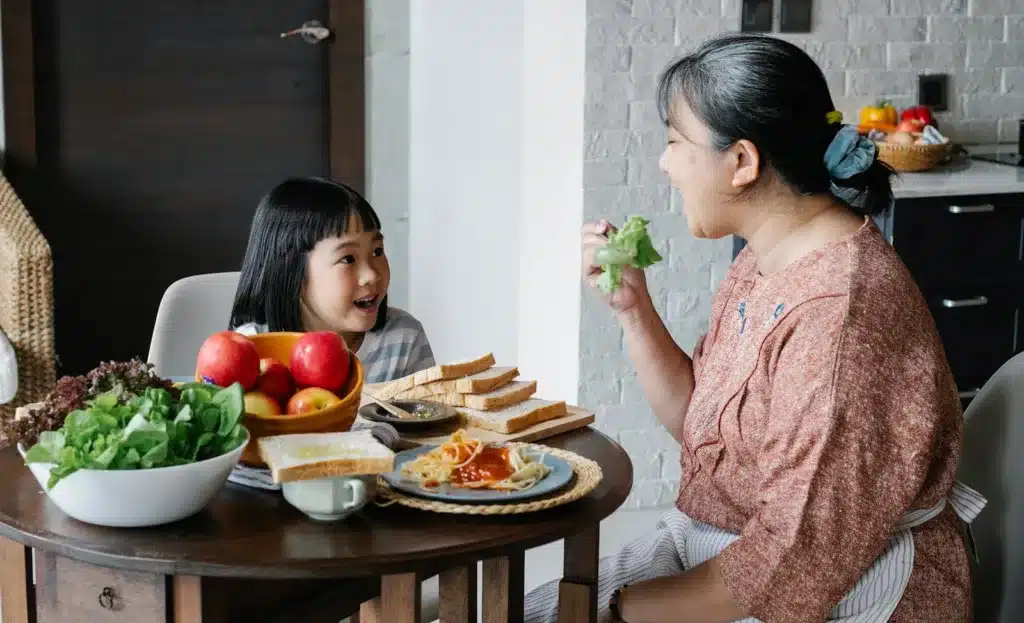 Side view of aged positive Asian woman in apron and dress eating lettuce while having lunch with adorable attentive little granddaughter sitting at table with opened mouth. mindful consumption, nutritional well-being