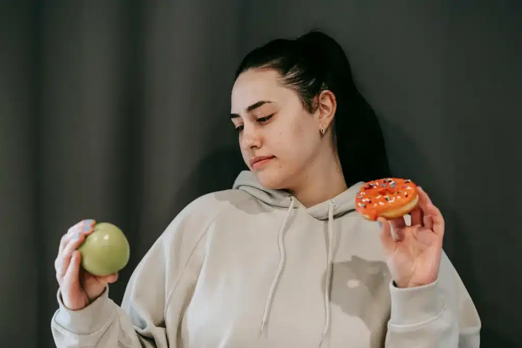 Pensive female wearing casual hoodie deciding between healthy green apple and sweet doughnut in studio. mindful consumption, nutritional well-being