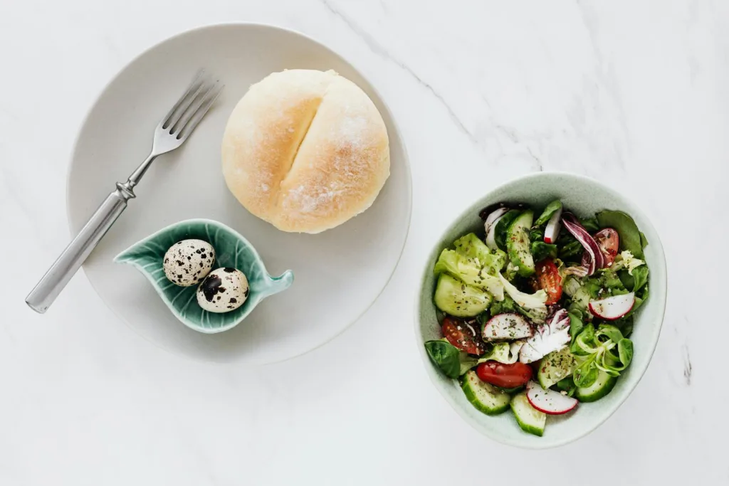 From above of healthy diet salad in bowl and plate with quail eggs fork and bun placed on white marble table, heart-healthy superfood recipes, organic food benefits, healthier life