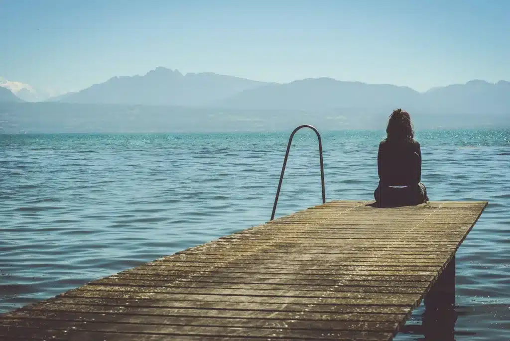 a woman sits on the end of a dock during daytime staring across a lake