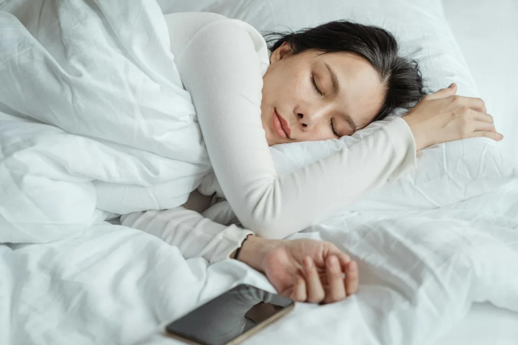 Calm Asian female wearing white pajama sleeping in comfortable bed with white sheets near modern mobile phone in morning. sleep quality, mental well-being, consistent sleep schedule, overall well-being